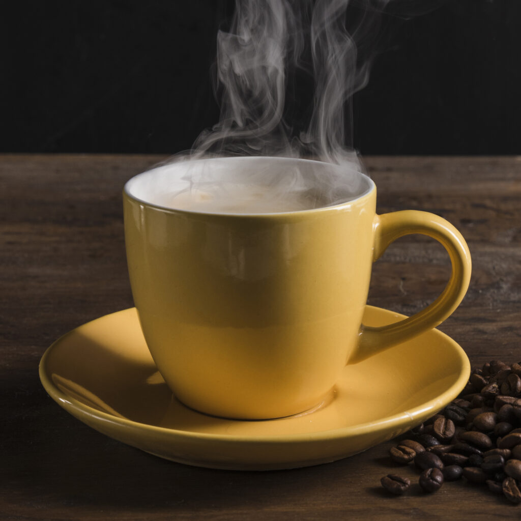 A cup filled with hot coffee next to scattered coffee beans on a wooden surface.