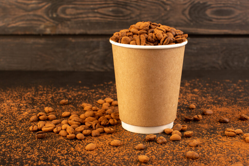 Brown roasted coffee beans filled inside a paper cup placed on a brown textured surface.